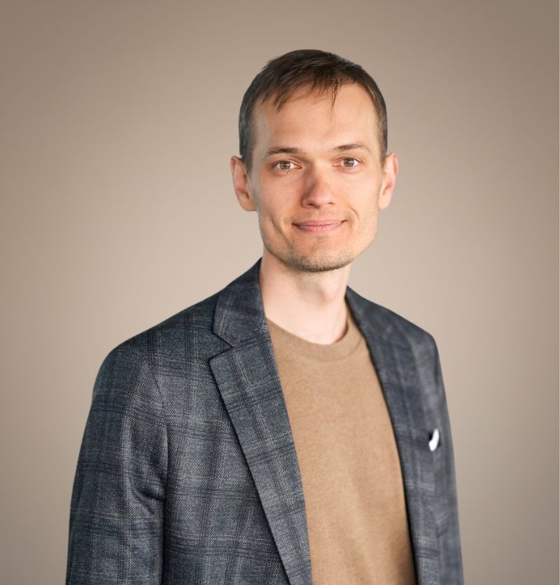 A confident person with short, styled hair and a leather jacket poses against a dark backdrop.