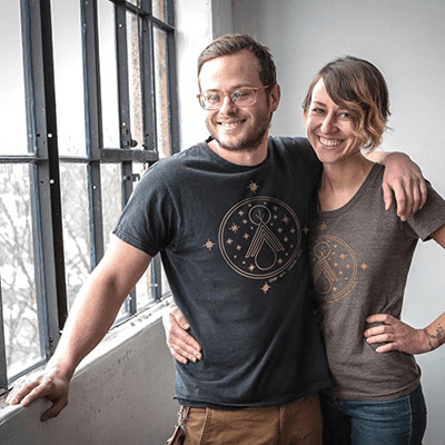 A man and woman stand smiling near a window. They are wearing matching t-shirts with a small design. The woman has her arm around the man.