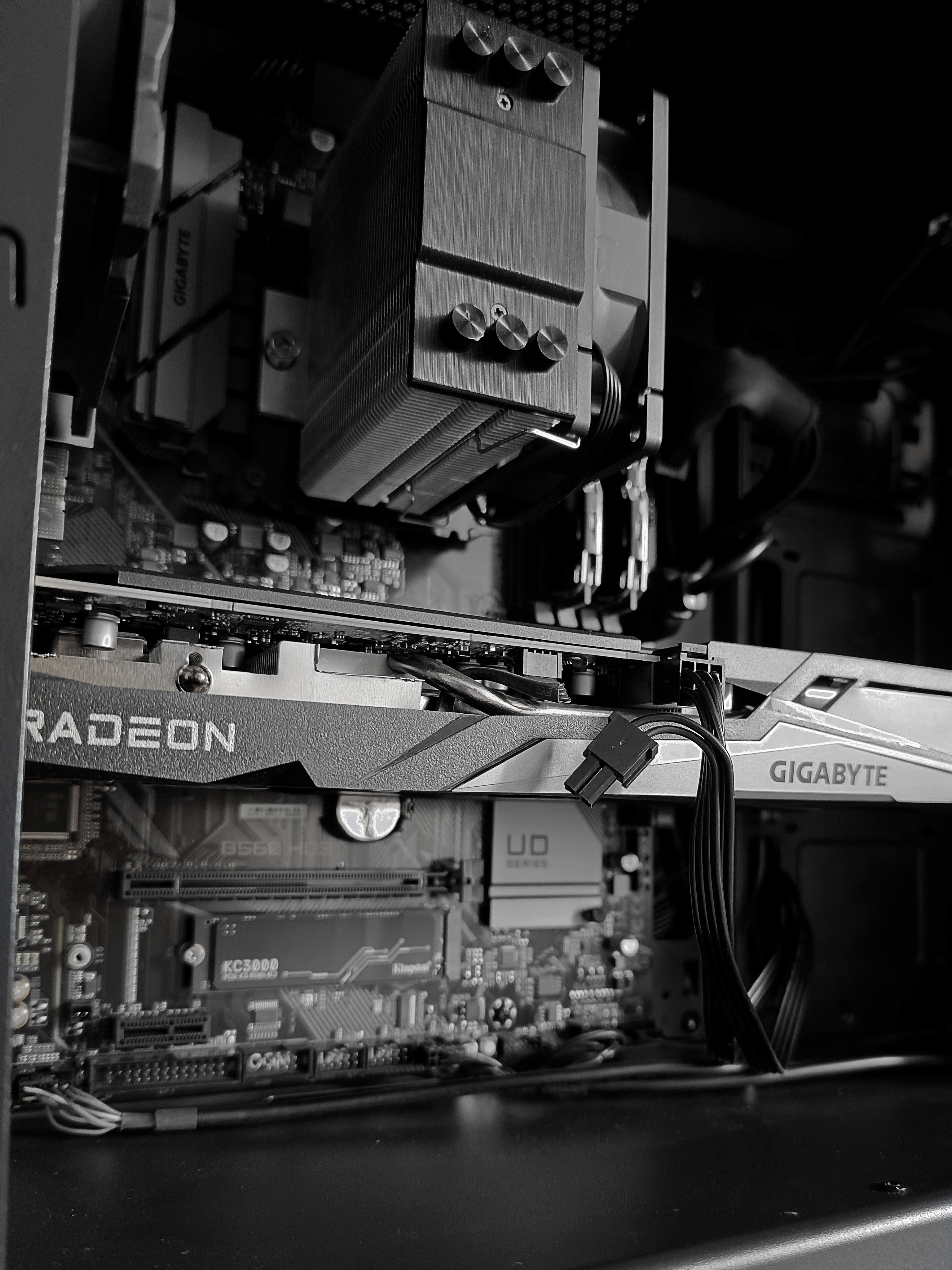 Czarno-białe zdjęcie wnętrza komputera z bliska. W centralnej części widoczna jest karta graficzna Gigabyte Radeon, a poniżej dysk Kingston KC3000 zamontowany na płycie głównej Gigabyte z serii UD. U góry znajduje się masywne, wieżowe chłodzenie procesora.