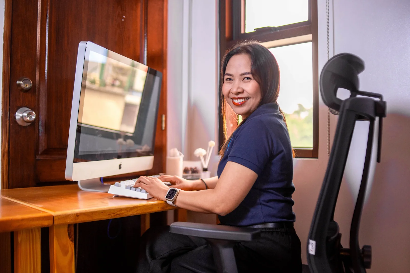 A woman turns and smiles at the camera while working on a desktop computer in a wooden office nook.