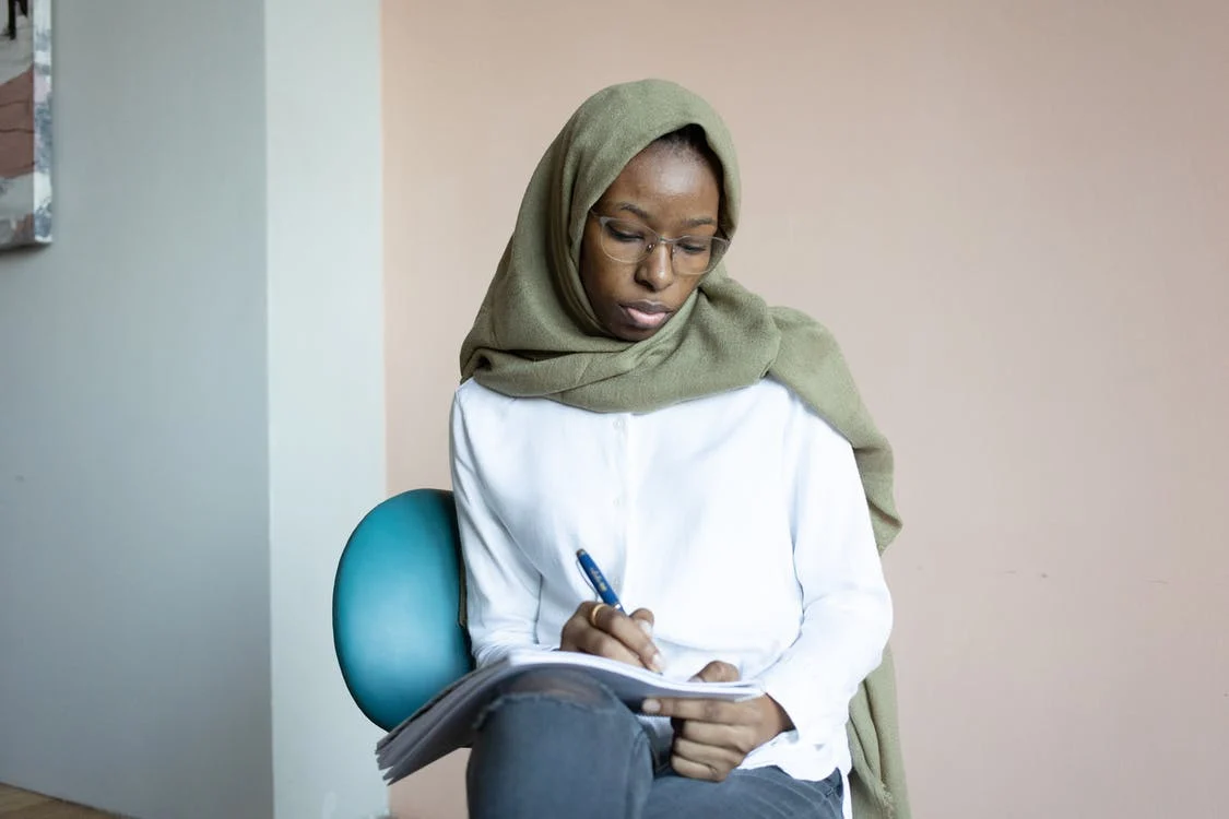 An image showing a women wearing a green scarf writing in a book