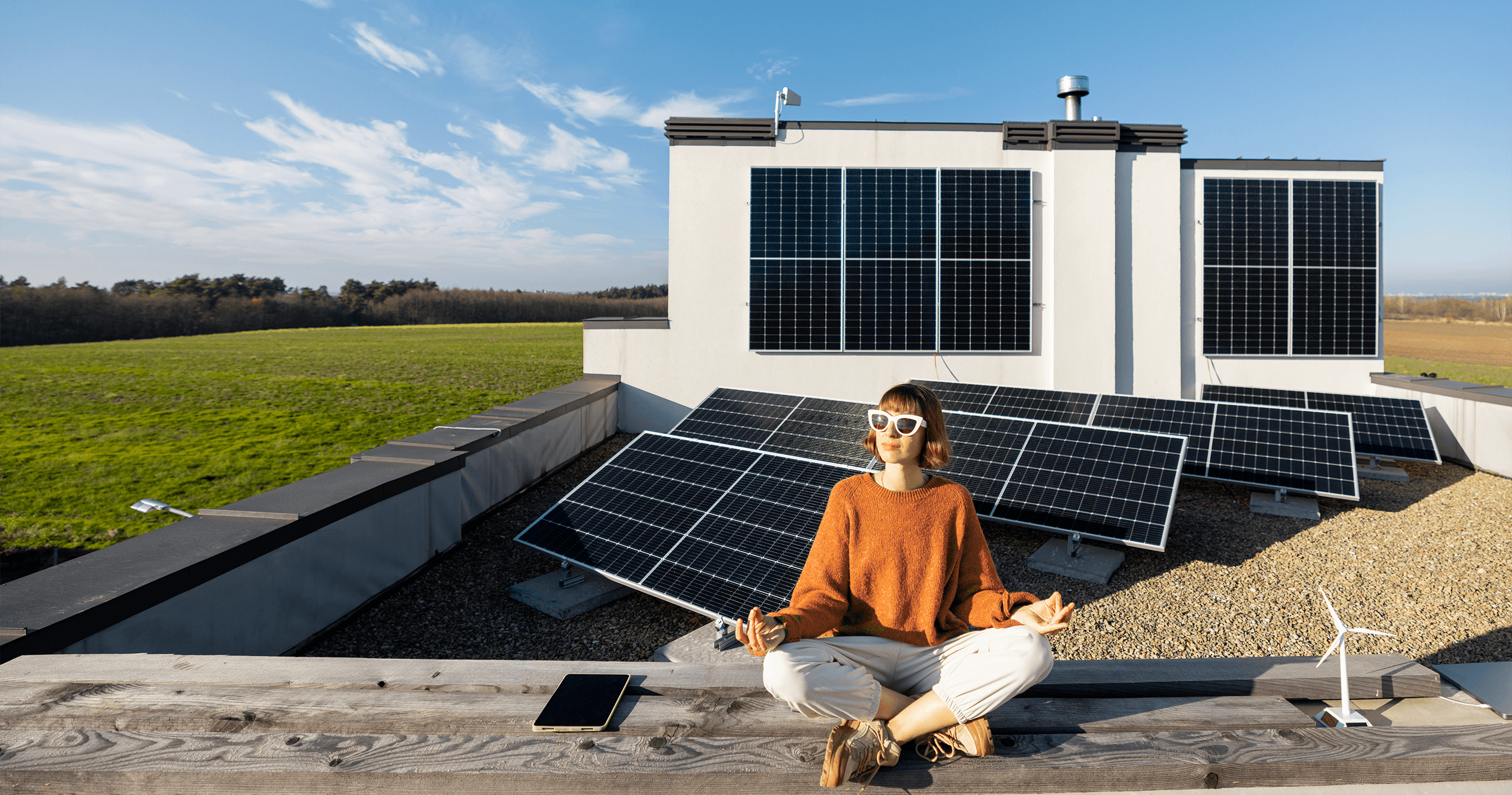 Eine Person sitzt mit gekreuzten Beinen in einer meditativen Haltung auf einem Dach, umgeben von mehreren großen Sonnenkollektoren, unter einem klaren blauen Himmel, wodurch erneuerbare Energien und nachhaltiges Leben hervorgehoben werden.