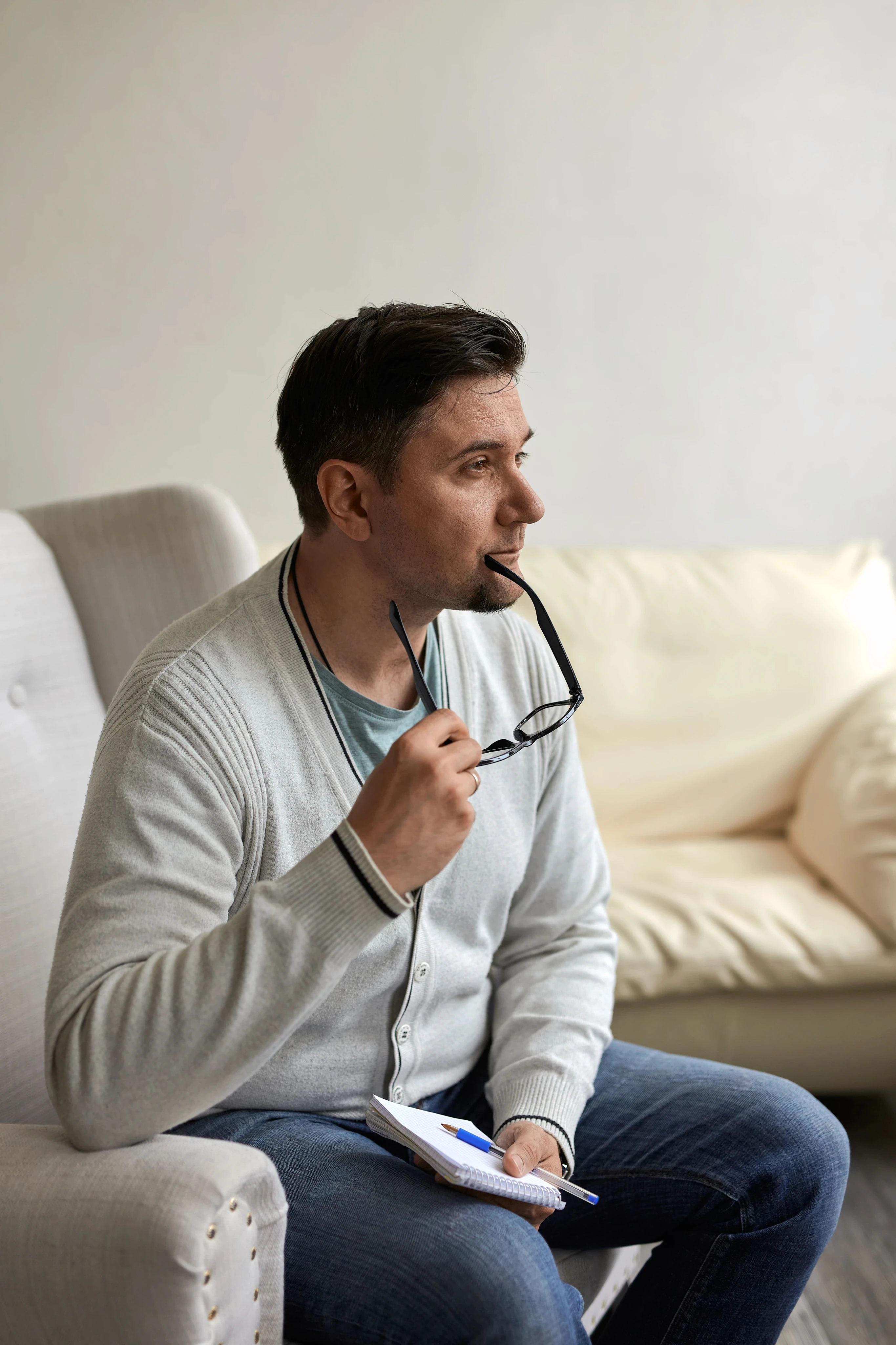 A man sits pensively in an armchair, holding glasses to his lips and a notebook in hand. He wears a light sweater and jeans, with a neutral room setting.