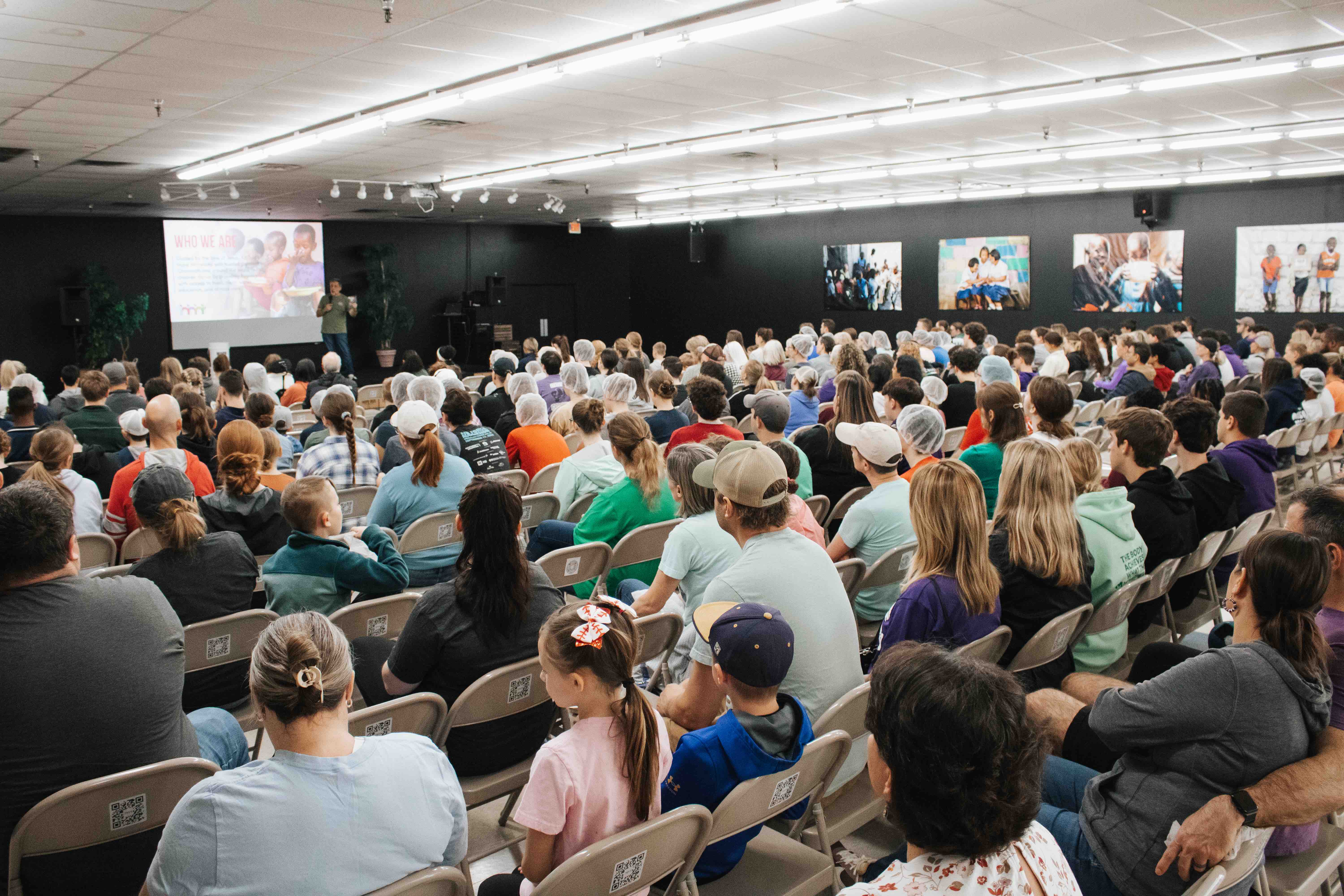 A large group of volunteers seated in rows, listening to a presentation at A Child’s Hope Int’l Hands Against Hunger™ packing event; the screen displays information about the organization.