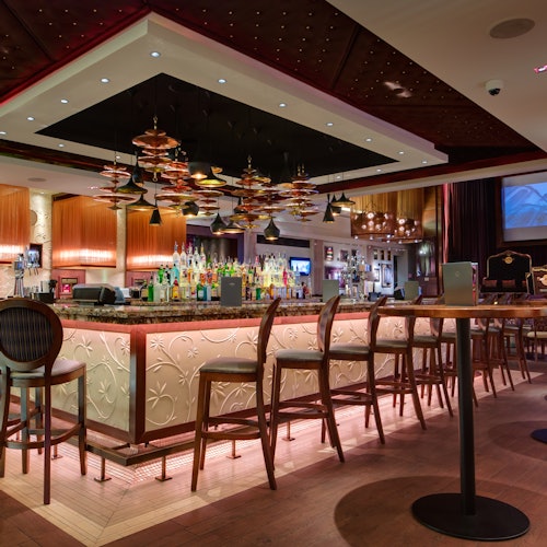 Modern bar with a central counter, high chairs, hanging light fixtures, and shelves of various bottles. Tables and more seating in the background.
