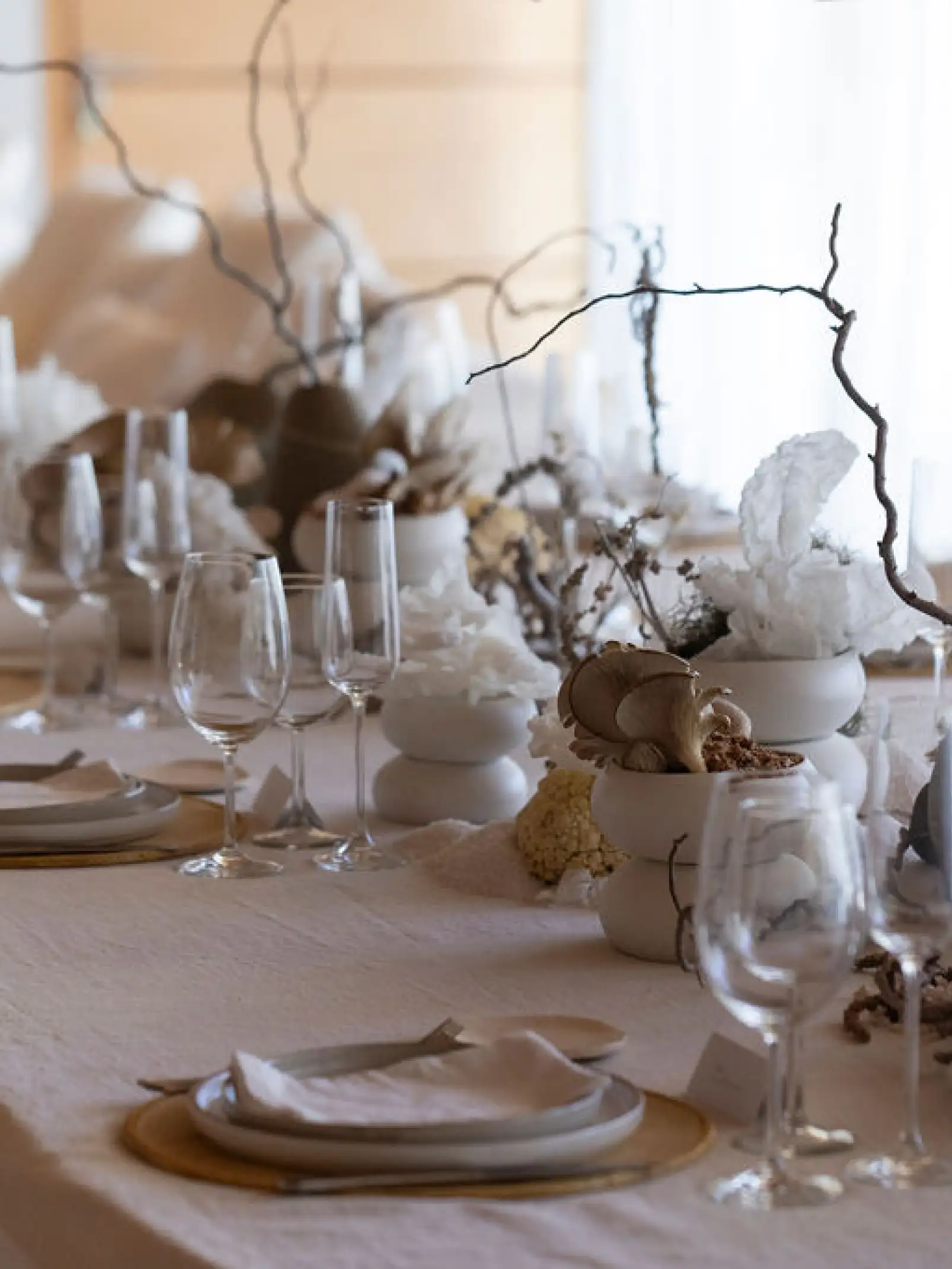Detalle de mesa elegante con vajilla minimalista, copas de cristal y centros decorativos de hongos, ramas secas y elementos blancos orgánicos.