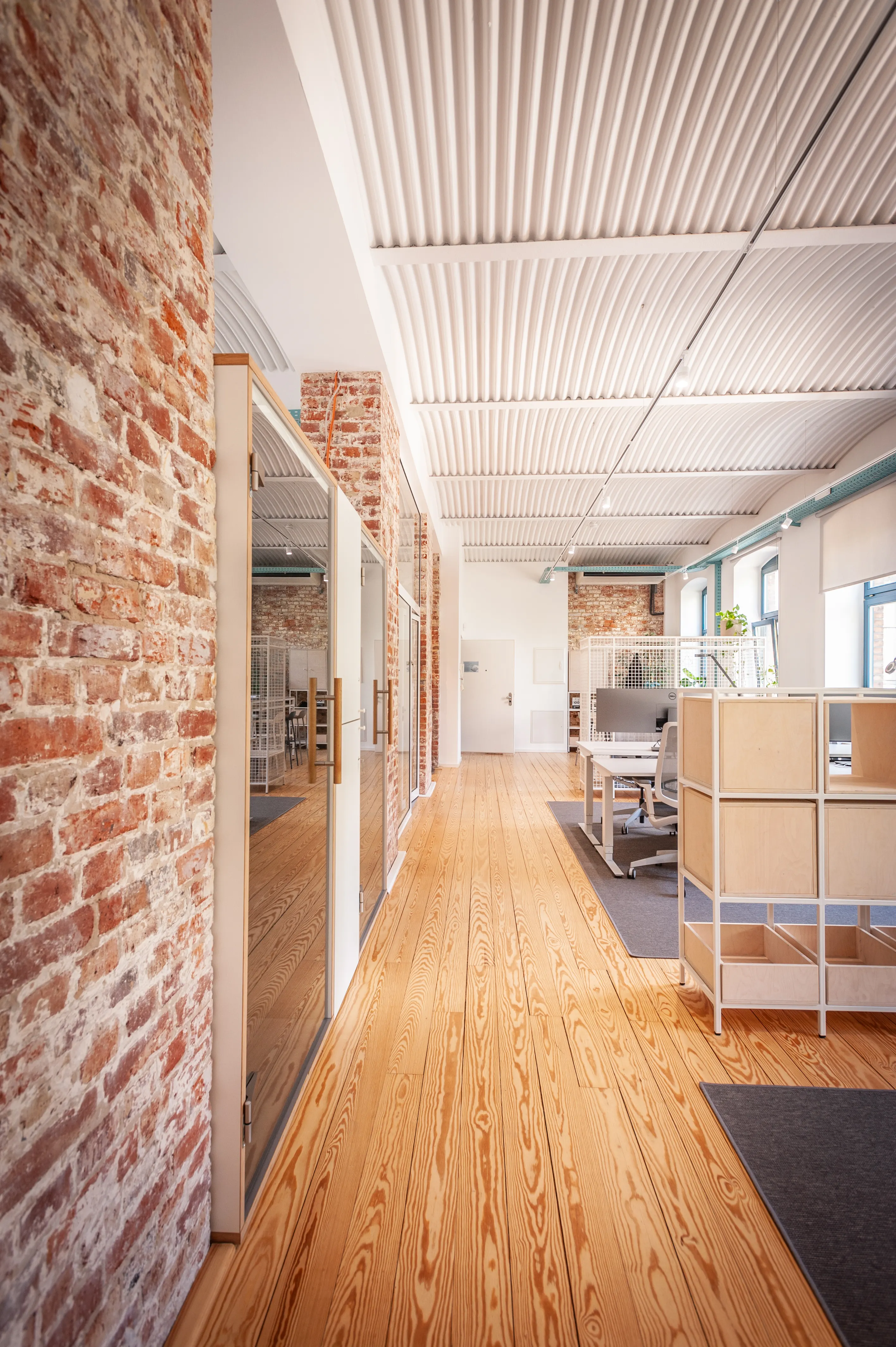 Corridor at EHEALTH-TEC with exposed brick walls, wood-framed glass office doors, pine plank flooring, a white corrugated ceiling and birch shelving units in the open office beyond.