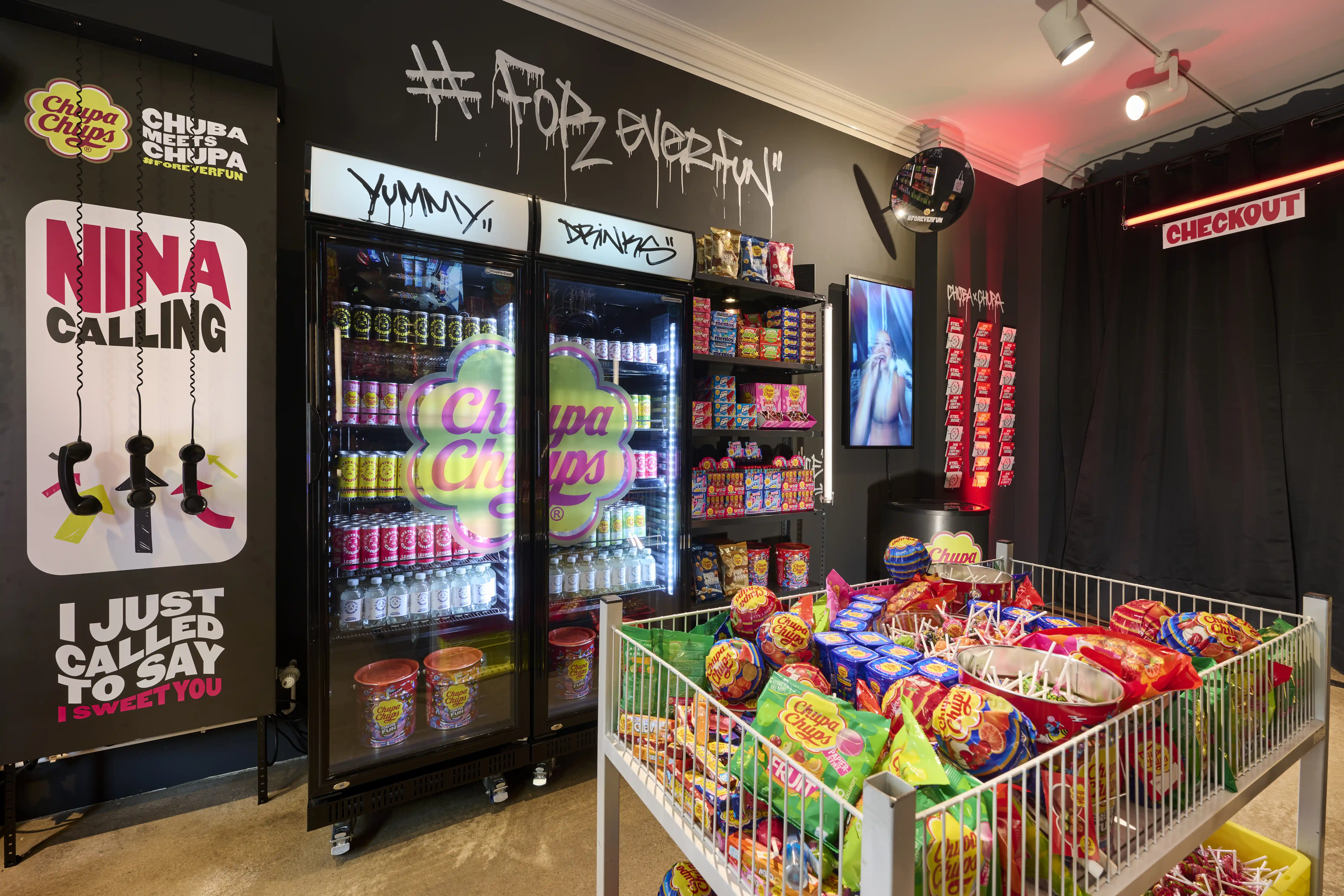 Interior of the Chupa Chups kiosk with black graffiti walls, drinks fridge and white wire cart full of candy.