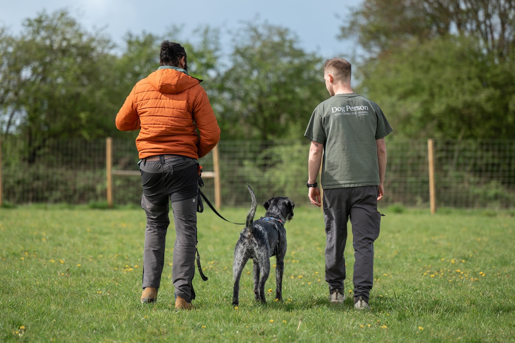 A happy, relaxed dog is between their owner & Scott (Dog Person). All three are walking away from the camera, where the lead is loose, and Scott is wearing a Dog Person branded t-shirt