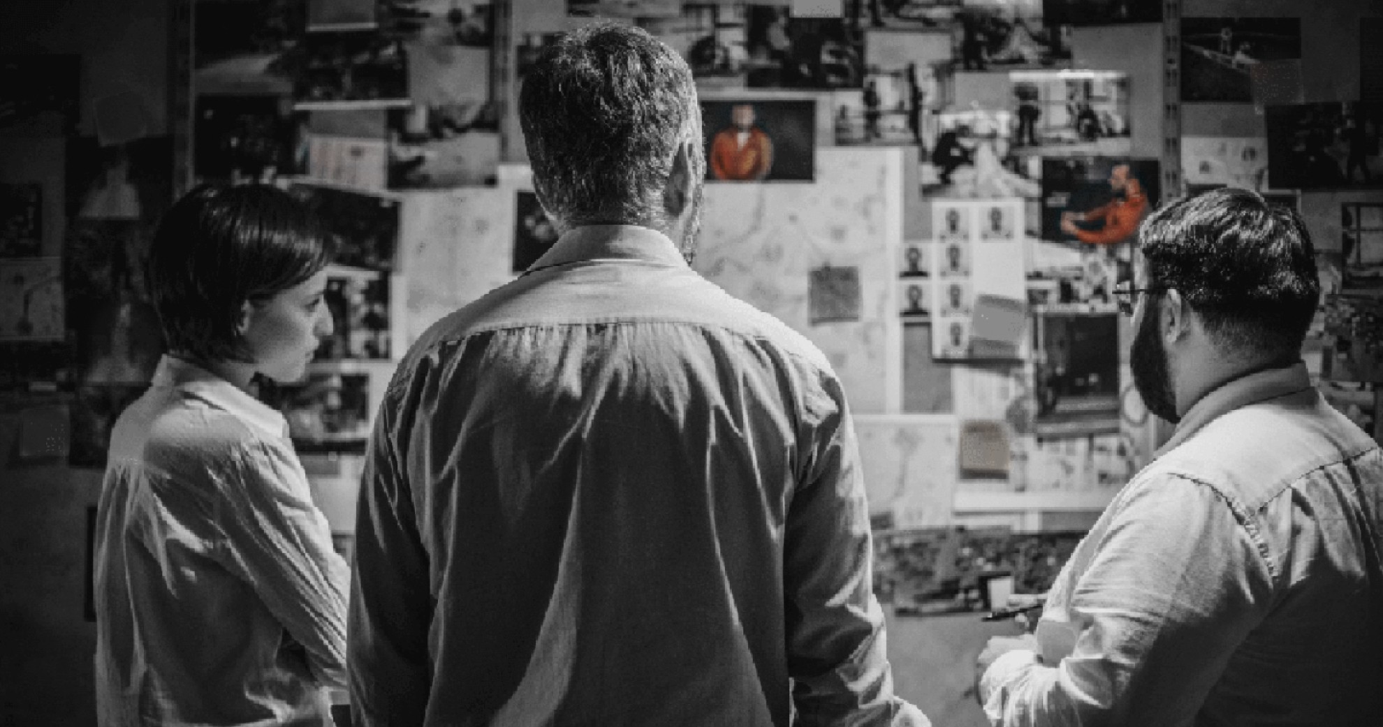 Black and white photo of 3 investigators discussing in front of a panel with clues.