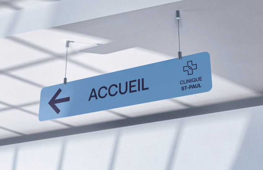 Panneau bleu suspendu dans un batiment en martinique avec inscription noire « ACCUEIL » et flèche vers la gauche, indiquant la réception de la Clinique St-Paul. Intérieur moderne et lumineux.