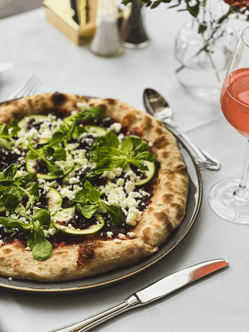A gourmet pizza topped with fresh spinach, sliced figs, crumbled feta cheese, and a sprinkle of herbs sits on a rustic brown plate next to a glass of rosé wine on a white tablecloth, creating an elegant dining setting.