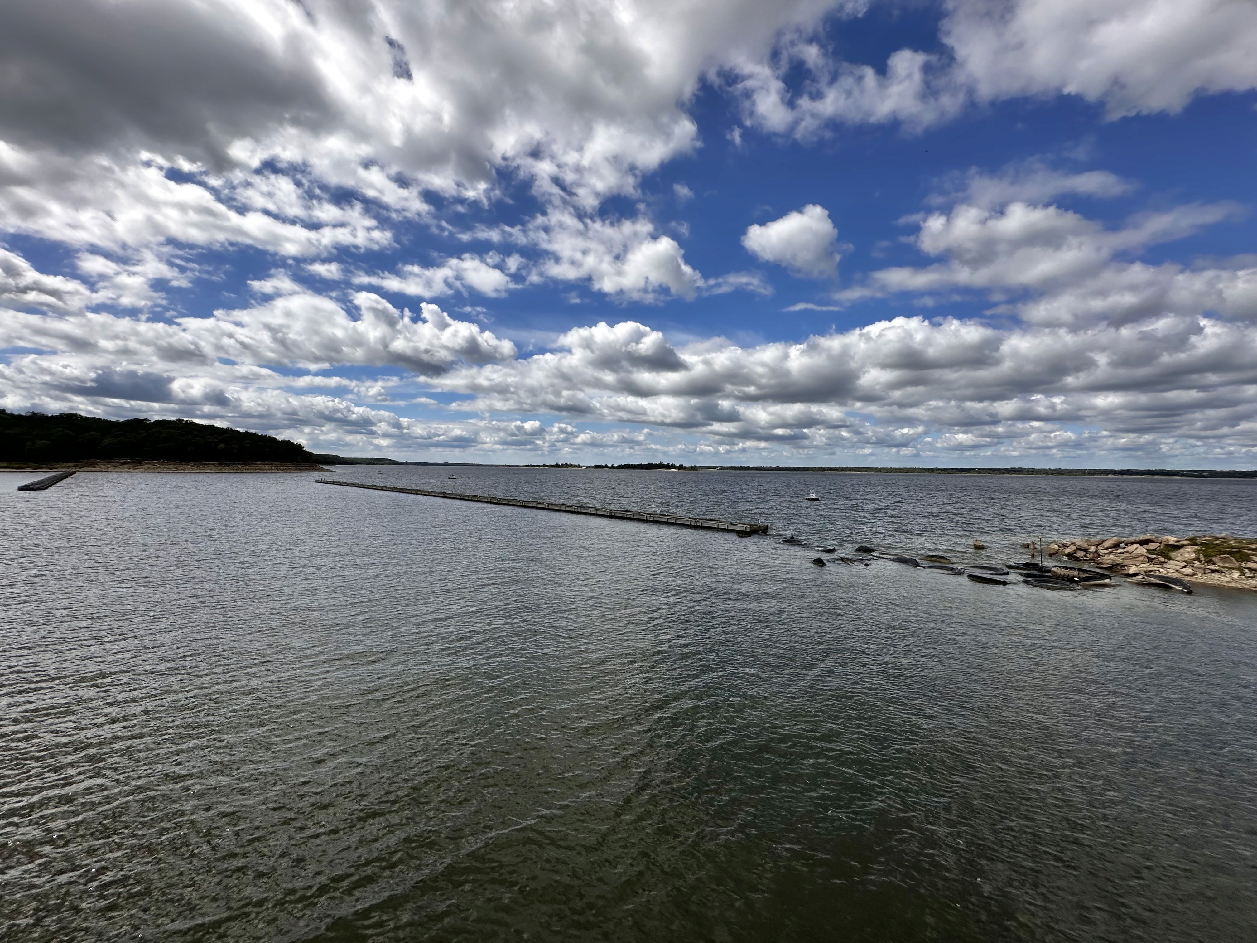 A serene coastal landscape featuring a vast, calm body of water meeting the horizon under a sky filled with scattered, fluffy clouds.