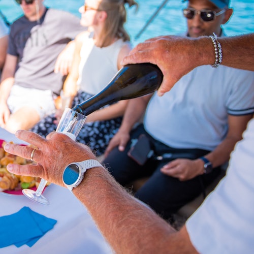 A person wearing a watch pours champagne into a glass on a boat, with three individuals seated in the background.