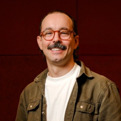 A young person with curly hair smiles at the camera while sitting in front of a laptop against a dark background.