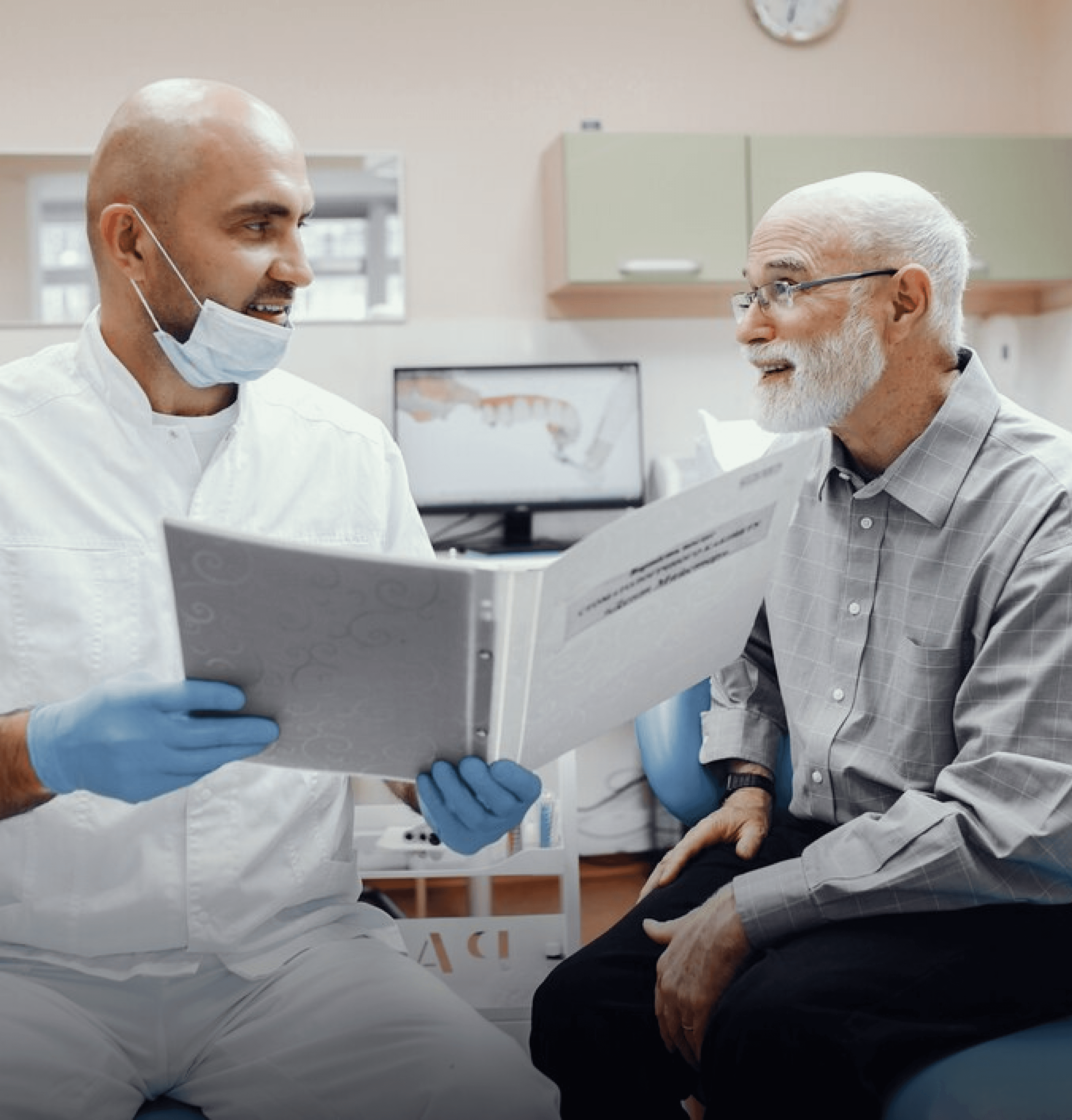 A dentist wearing a mask holding a brochure and discussing treatment options with an elderly male patient, both smiling and engaged in conversation.