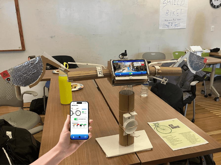 Overview of bike controls made of cardboard and a hand to the side holding a phone with the home screen of the mobile app