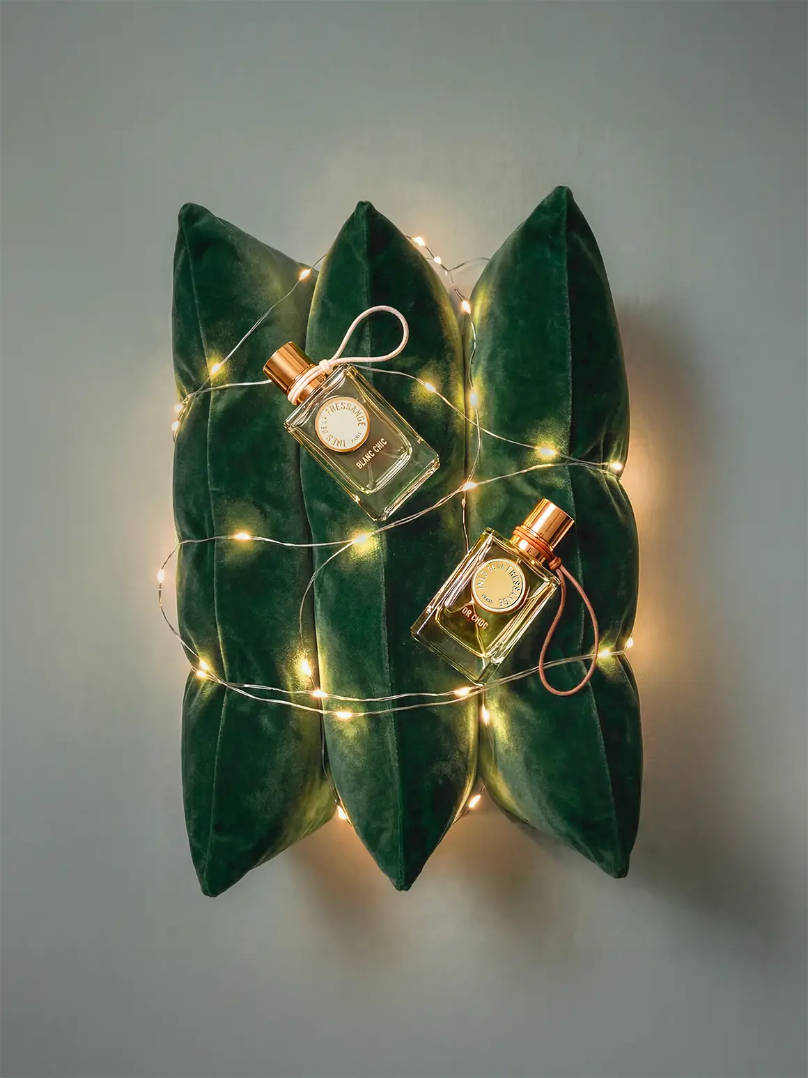 A pair of elegant glass perfume bottles with gold caps are gracefully placed on a cushion of dark green velvet, surrounded by soft, glowing fairy lights, creating a luxurious and high-end visual that aligns with a sophisticated fashion brand aesthetic.