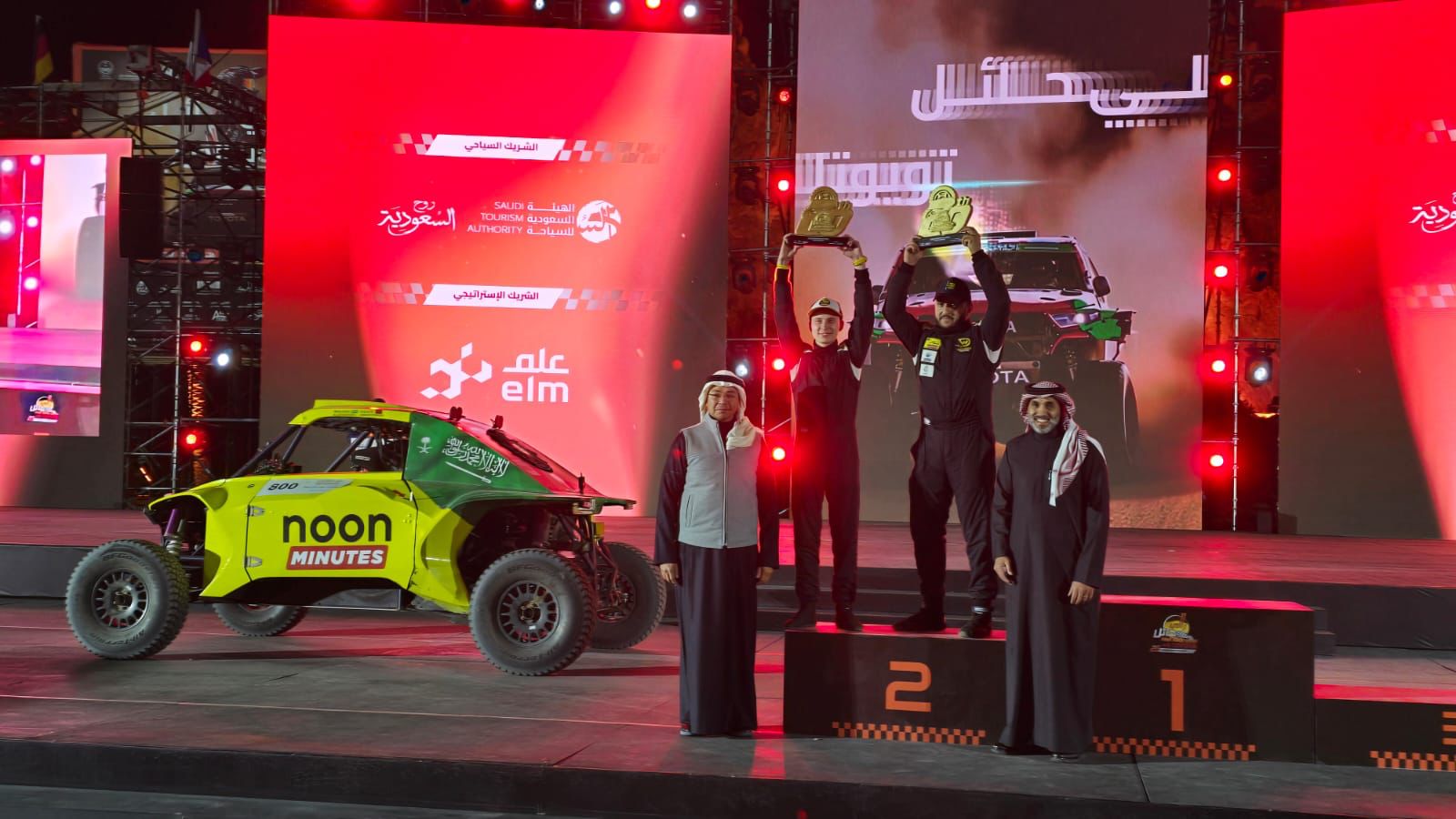 Three people stand on a podium holding trophies with two men in traditional attire nearby. A yellow and green racing vehicle is beside them. Bright stage lights and banners in the background.