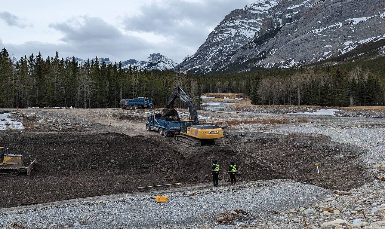 Excavation crew and heavy equipment removing accumulated sediment from Evan Thomas Creek sediment trap in Kananaskis with snow-covered Rocky Mountain peaks in background