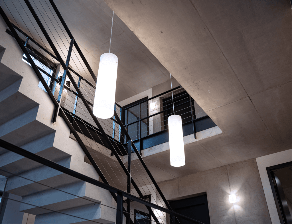 Architektonisches Innenraumfoto eines modernen Treppenhauses der kba Architekten und Ingenieure GmbH mit Sichtbeton, schwarzen Stahlgeländern und zylindrischen Pendelleuchten, die mehrere Ebenen verbinden.