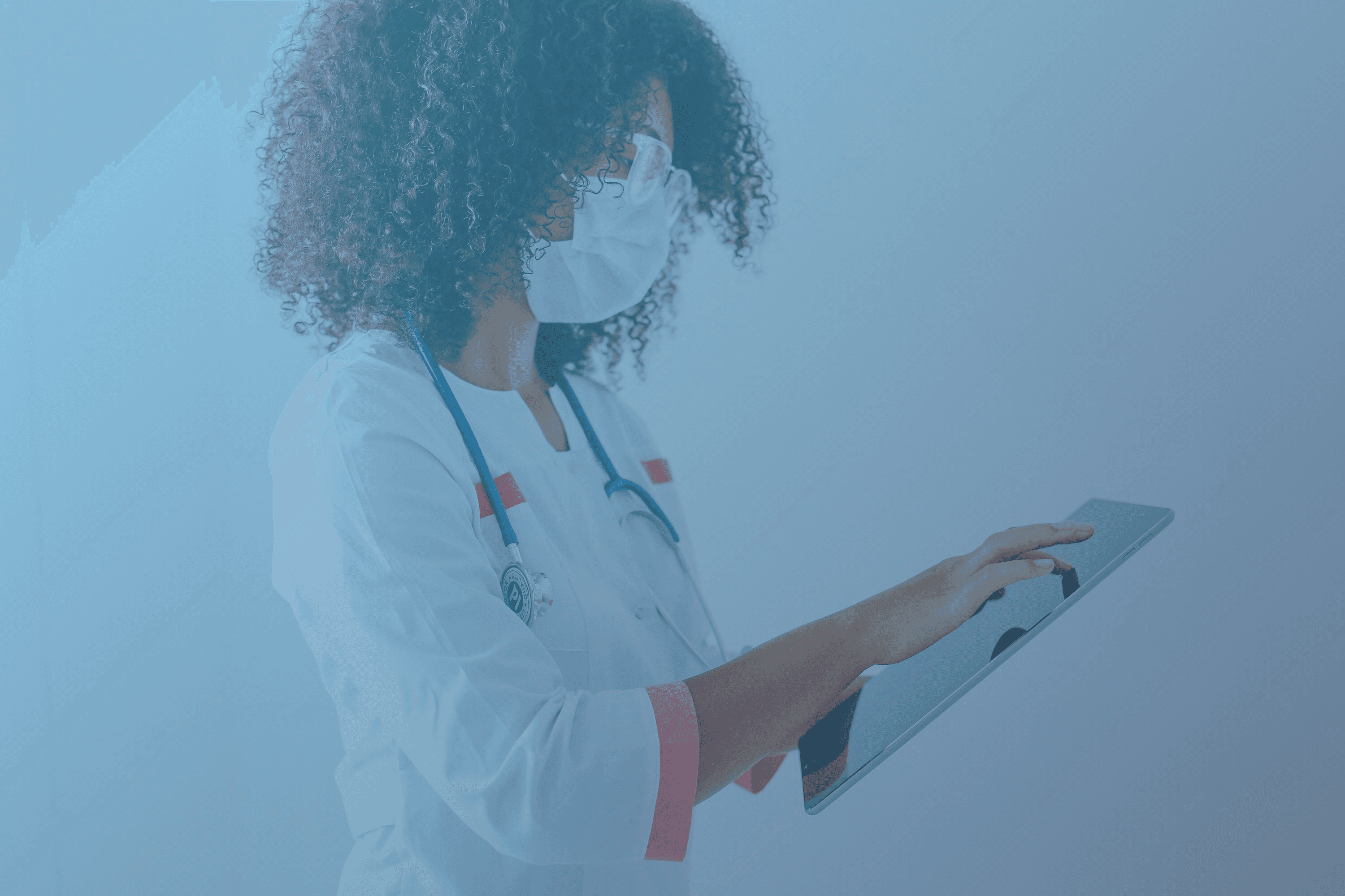 Eine Ärztin mit weißem Kittel, Stethoskop und Schutzmaske hält ein Tablet in der Hand und tippt darauf.