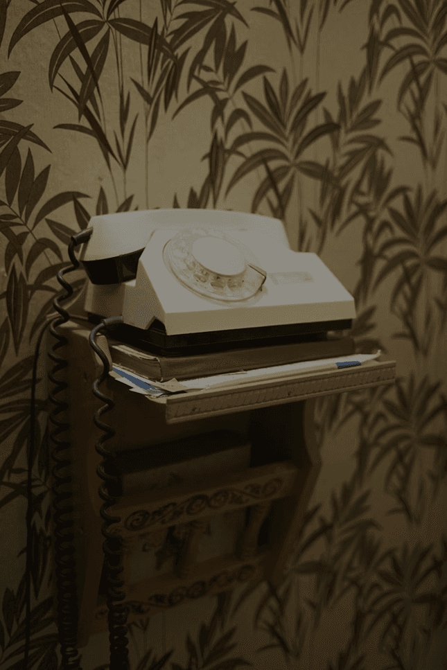 Vintage rotary telephone placed on a small wooden shelf, with papers stacked underneath and patterned wallpaper in the background.
