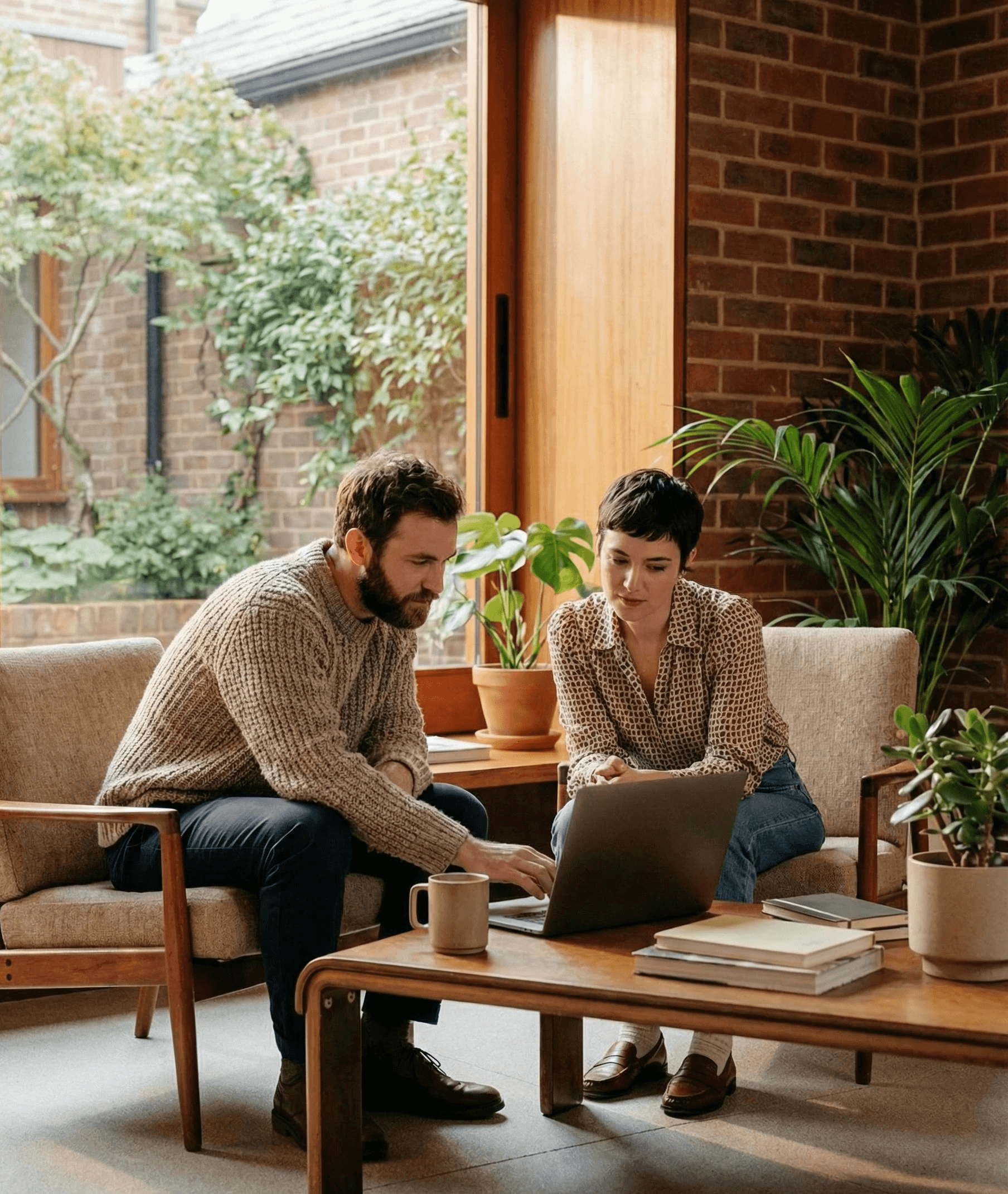 A man and woman sitting in armchairs, collaborating on a laptop in a cozy, plant-filled room.