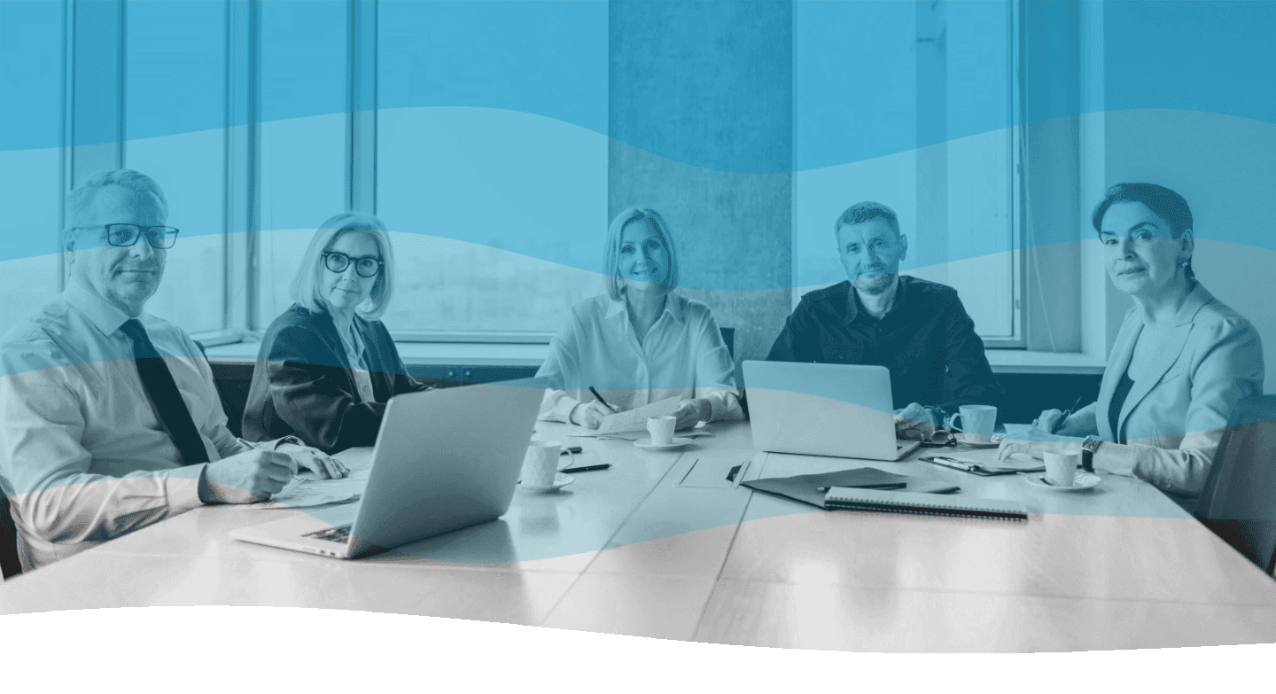 A group of professionals sitting at a boardroom table with open laptops, notepads, and coffee cups, engaging in a collaborative meeting.
