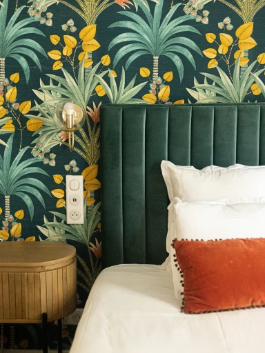 A stylish bedroom corner featuring an upholstered green velvet headboard, vibrant tropical-patterned wallpaper with palm leaves and berries, a round wooden bedside table, and a plush bed accented with a rust-colored cushion.