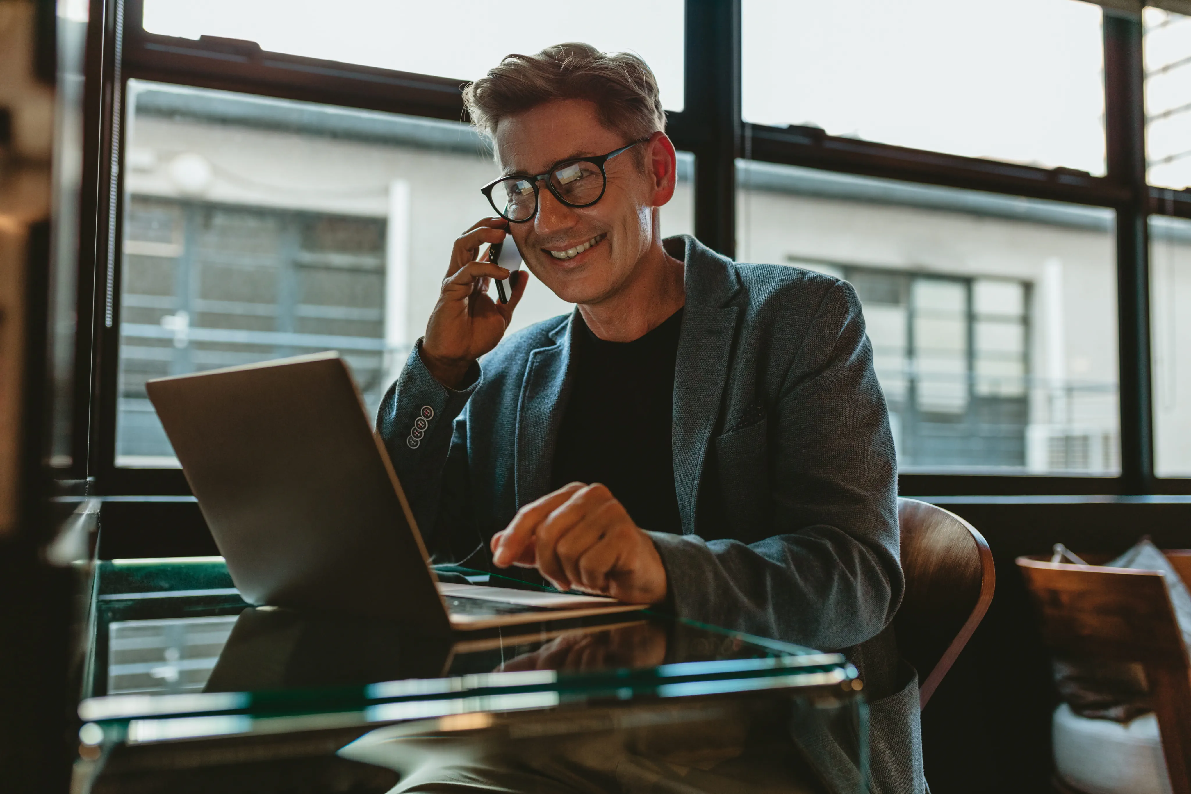 Older man in glasses sitting with laptop on coffee table, smiling while working.