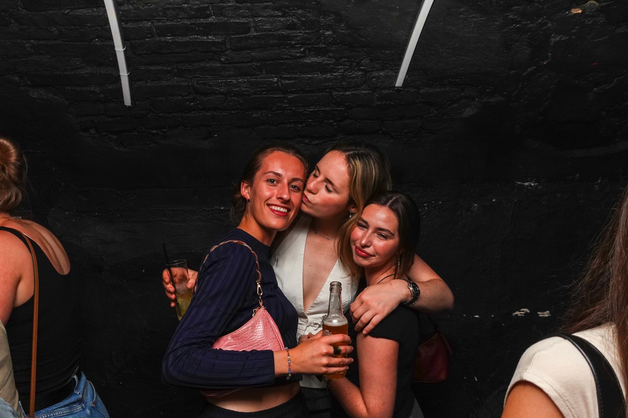Three young women posing closely together in an underground bar during a bar crawl in Nice holding drinks and smiling showing friendship spontaneity and the intimate social atmosphere of a lively nightlife experience on the French Riviera