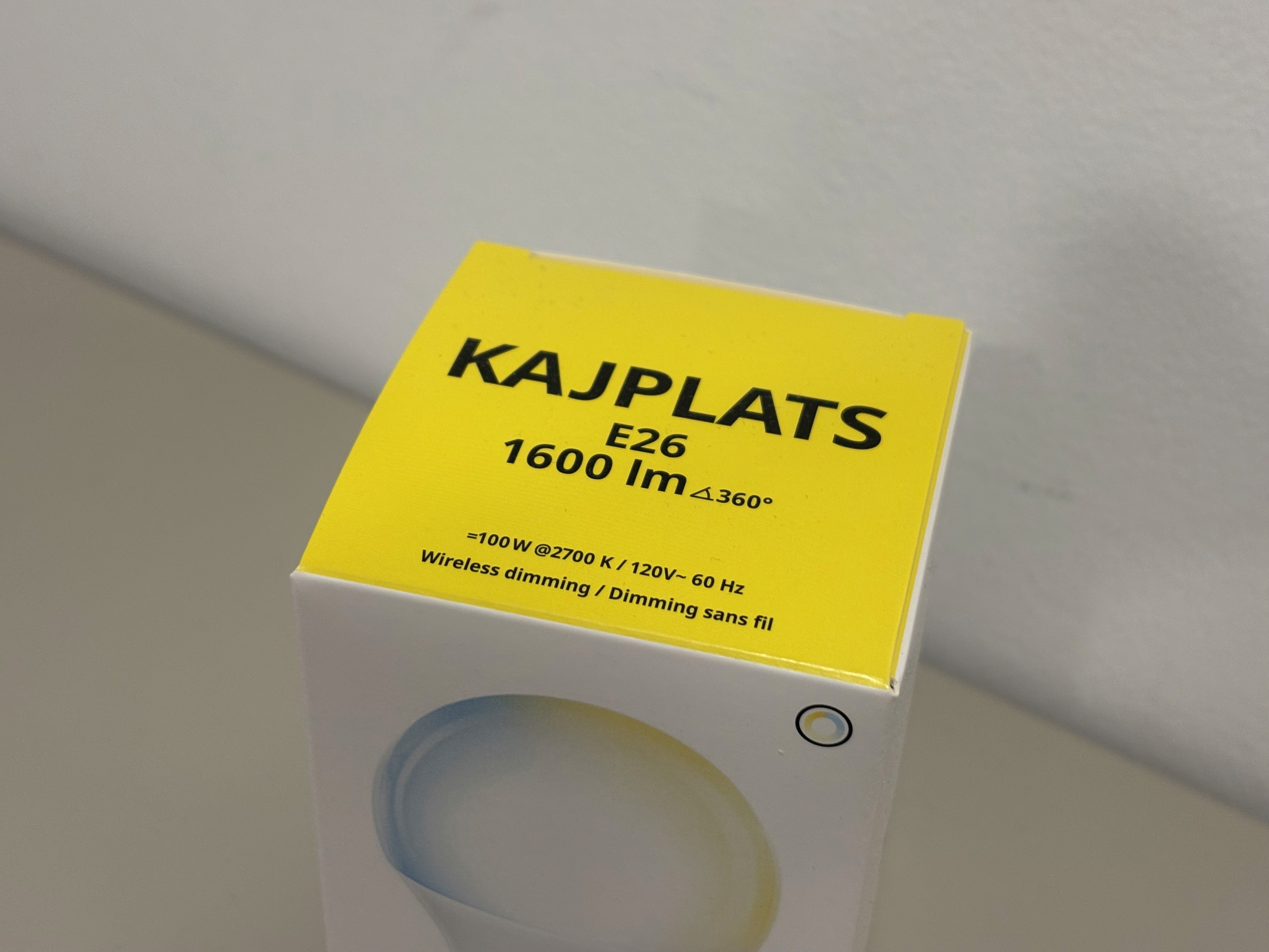A yellow and white box displays the label "KAJPLATS E26 1600 lm" for a dimmable 100-watt light bulb, placed on a light-colored surface.