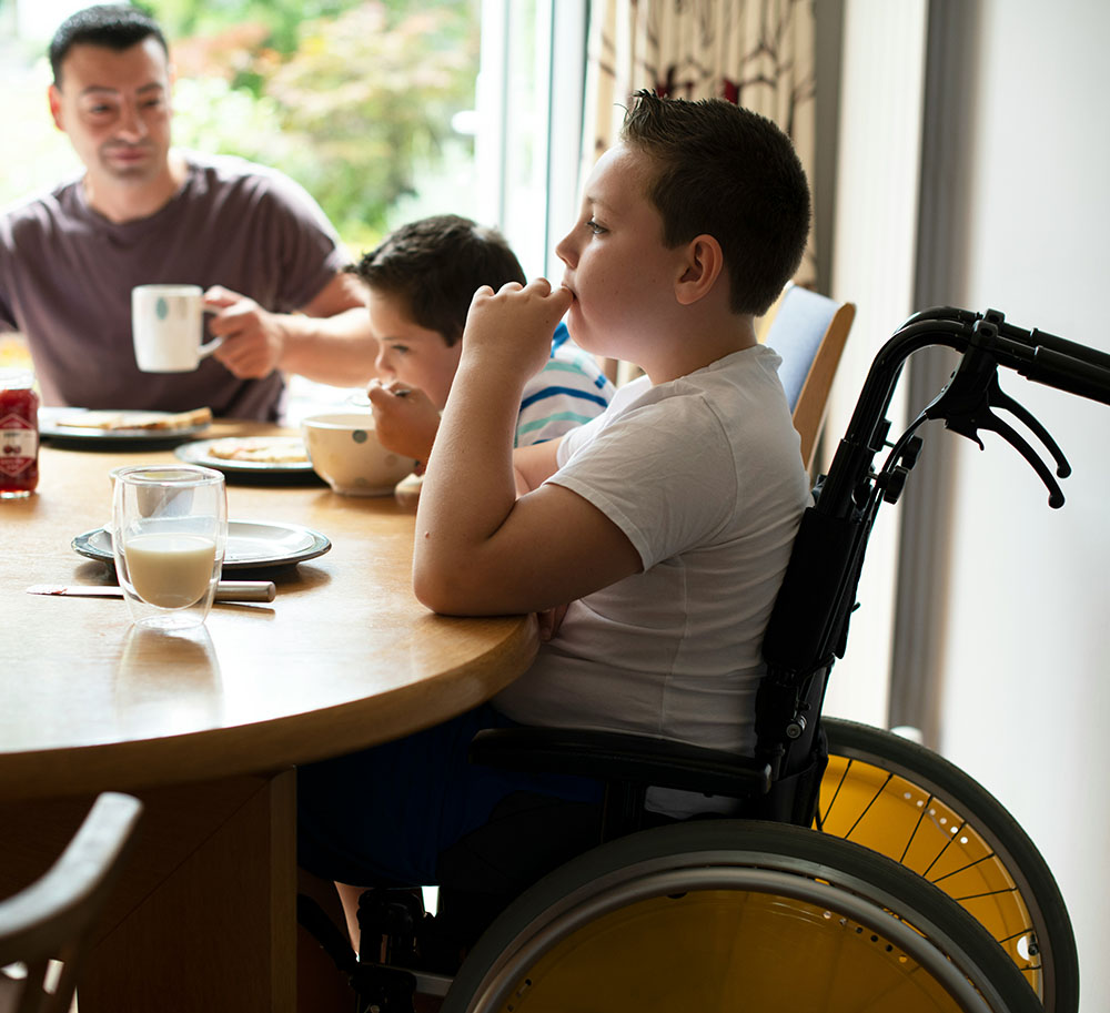 A young boy in a wheelchair sitting at a table with a man and another boy.