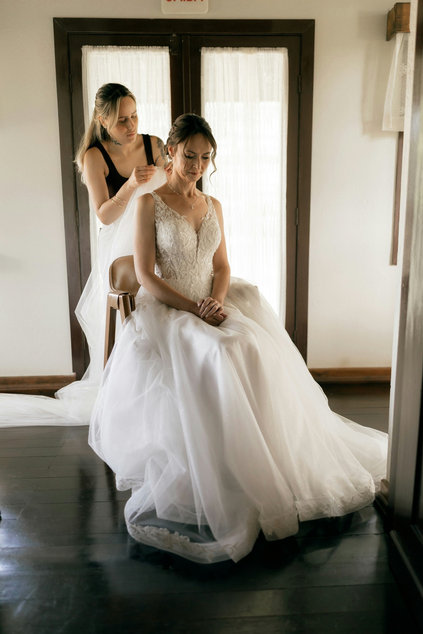 Bride seated in her wedding dress while an attendant adjusts her veil in a getting-ready room with soft natural window light