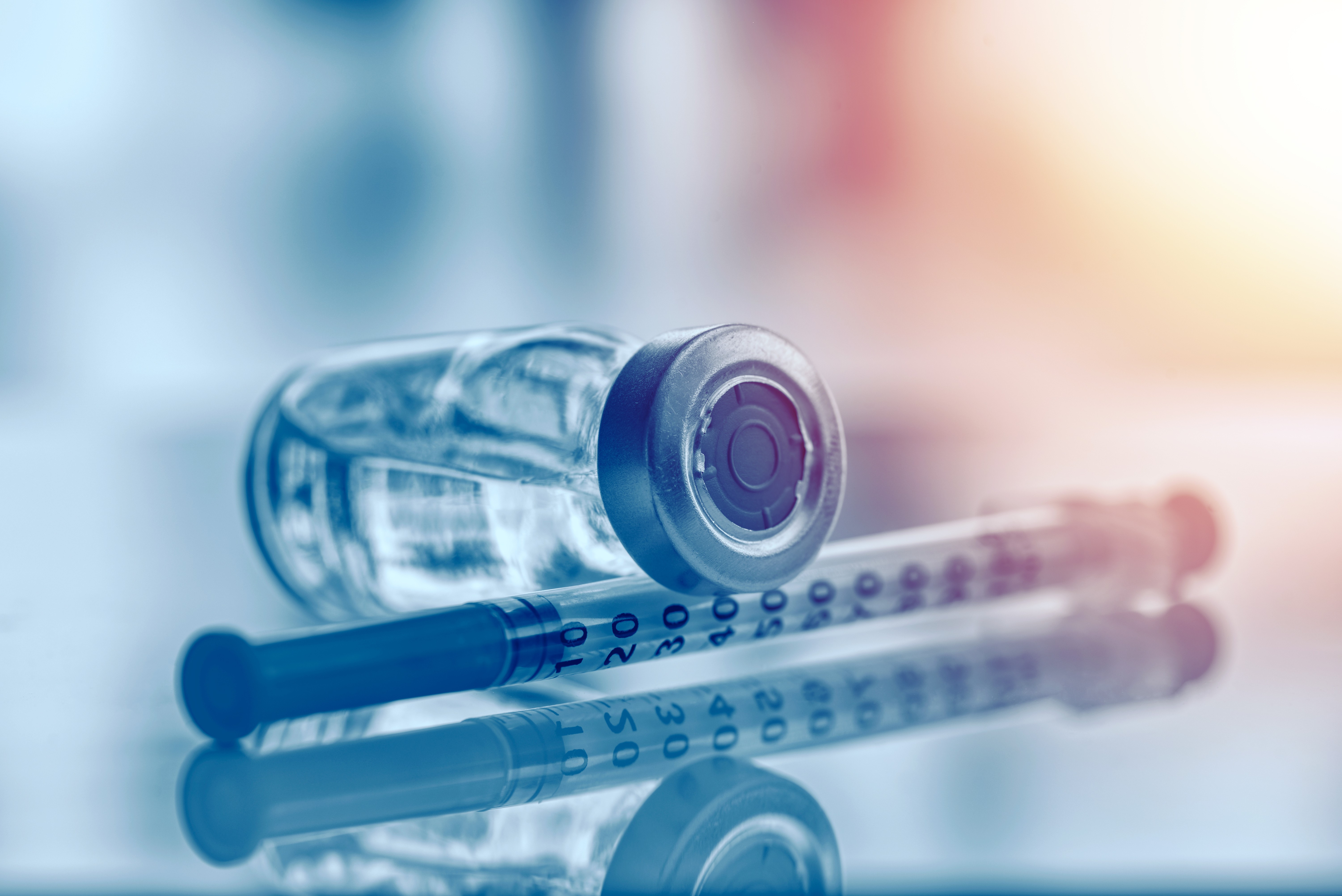 Close-up of a clear medicine vial lying beside two insulin syringes on a reflective surface, in a blue tone
