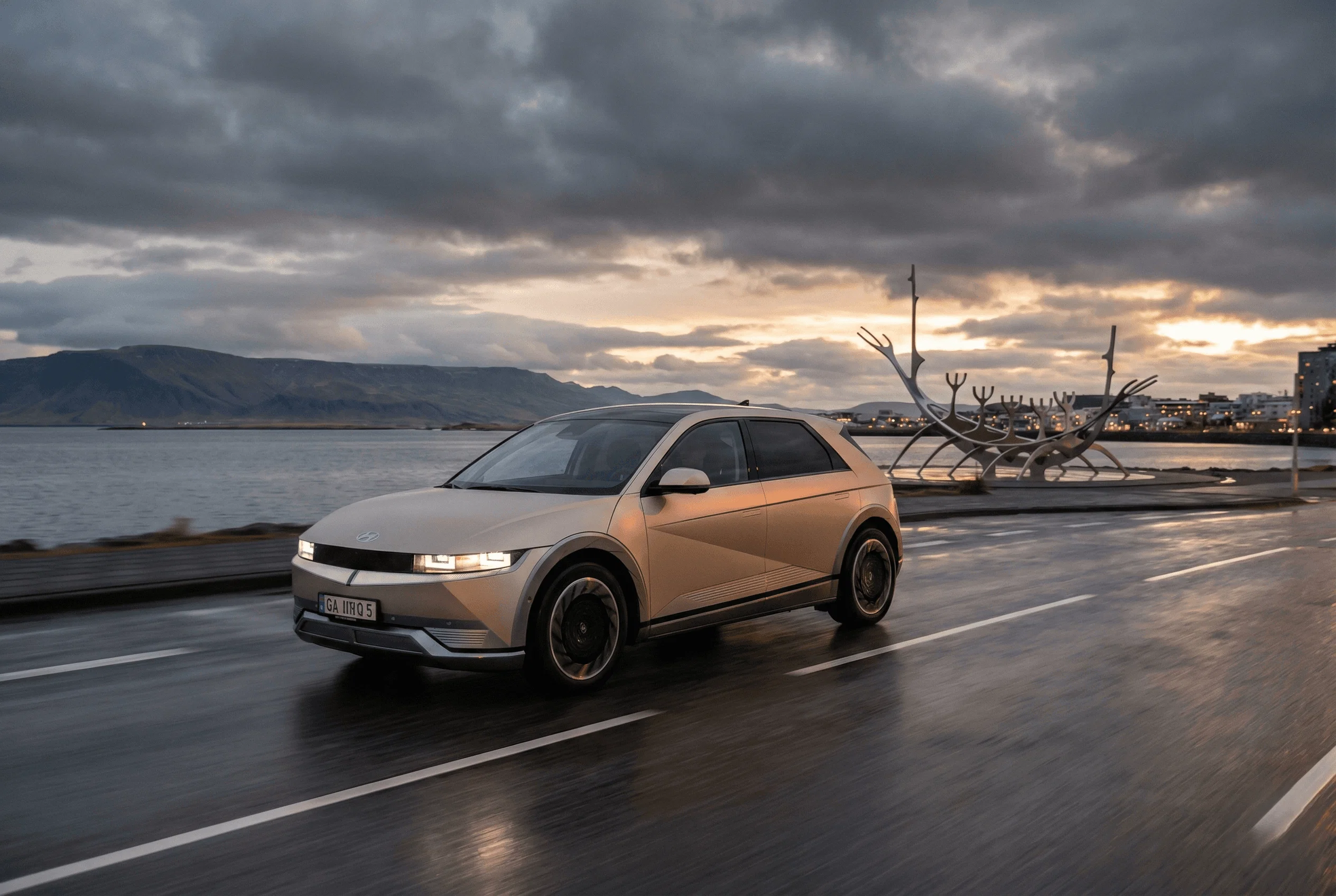 A beige electric SUV driving along a coastal road at sunset with mountains and the ocean in the background.