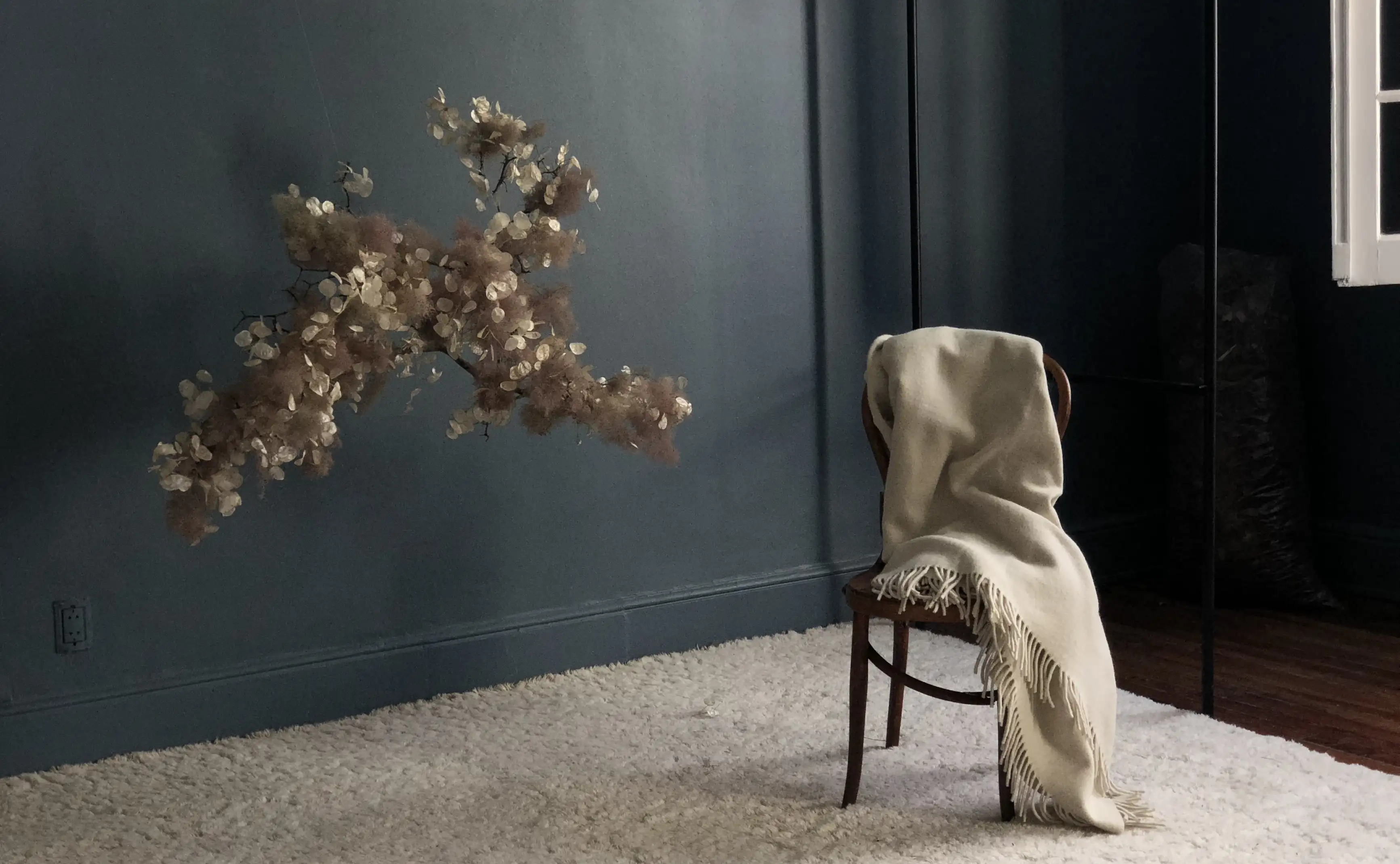 Composición minimalista con silla de madera, manta de lino y arreglo floral suspendido para diseño interior contemporáneo.