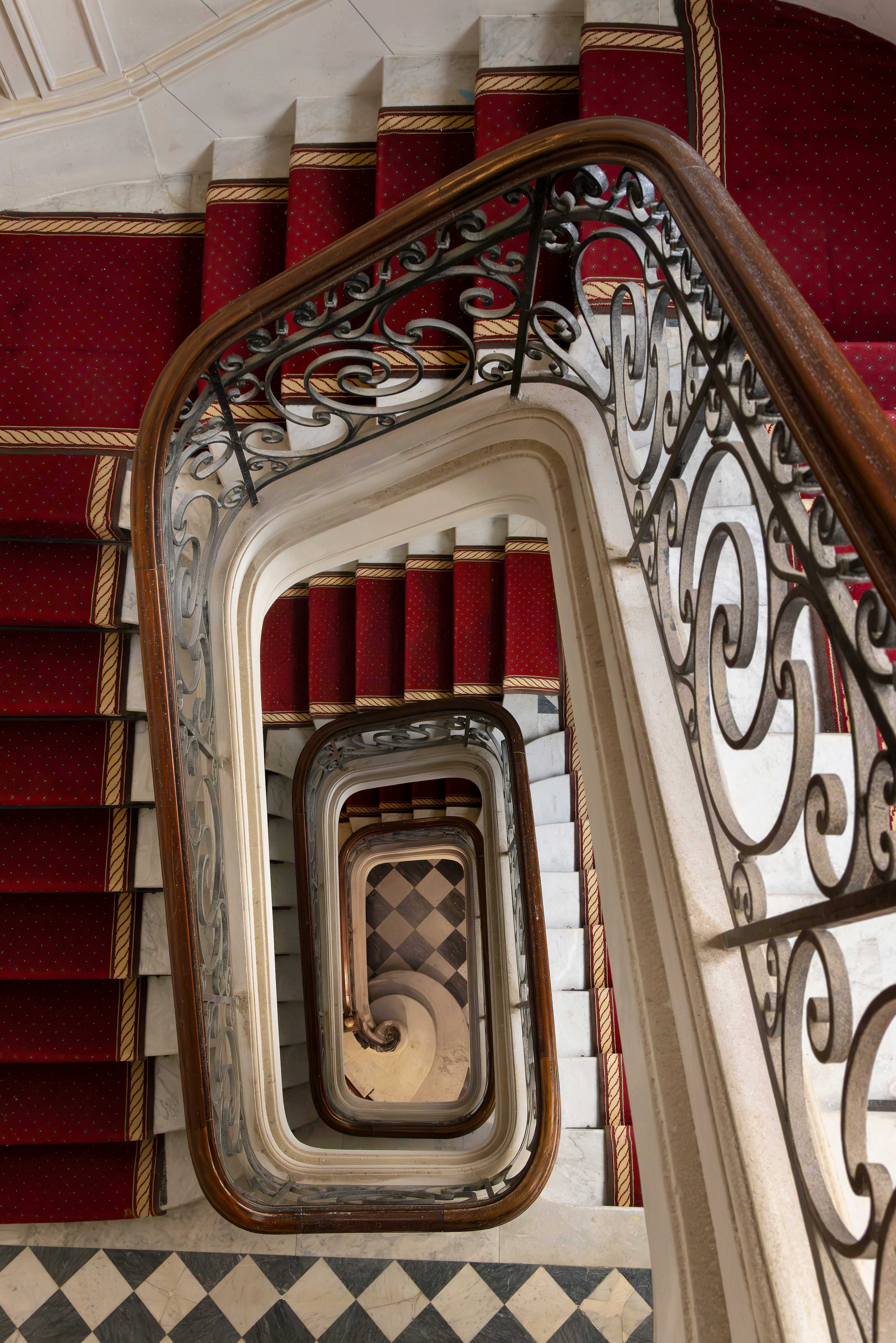 Vue plongeante de la cage d'escalier en spirale avec tapis rouge, rampe fer forge et sol en damier noir et blanc