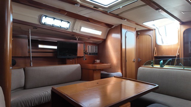 Dufour 40 Performance saloon interior looking aft, showing port and starboard grey upholstered settees flanking the polished cherry wood dining table, nav station with flat-screen TV and instrument panel, overhead hatch, companionway door open to the cockpit, and warm teak-and-holly sole flooring throughout