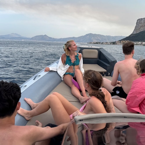A group of people in swimwear relaxes on a boat with mountains and a coastline in the background.