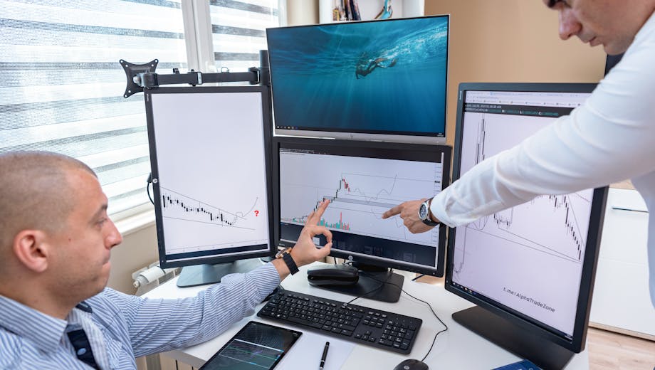 Two business professionals analyzing financial data on multiple computer screens in an office setting.