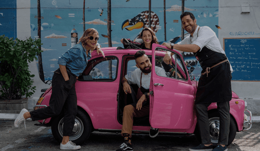 Four people smiling around a bright pink vintage car parked in front of a colorful mural, representing the Macchialina team.