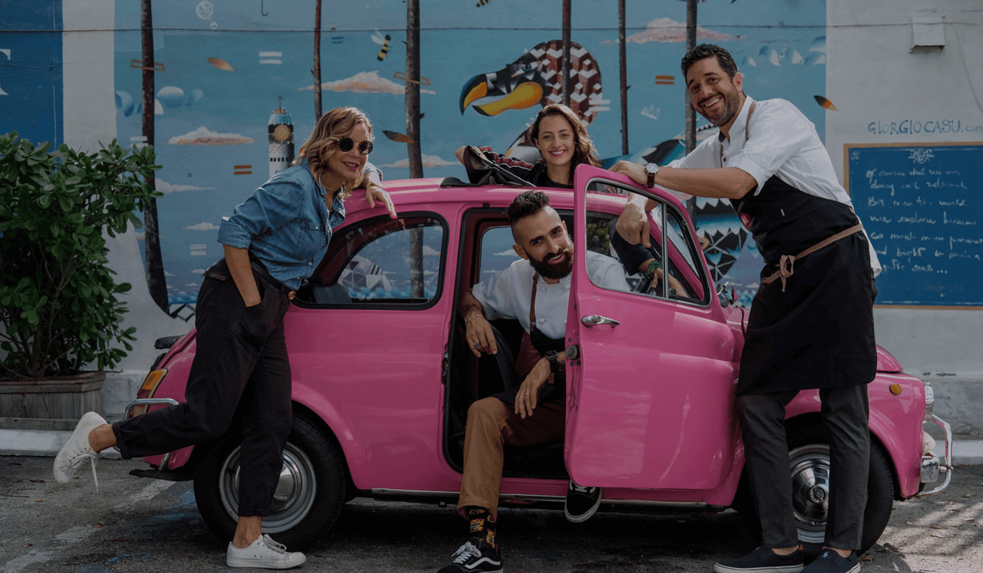 Four people smiling around a bright pink vintage car parked in front of a colorful mural, representing the Macchialina team.