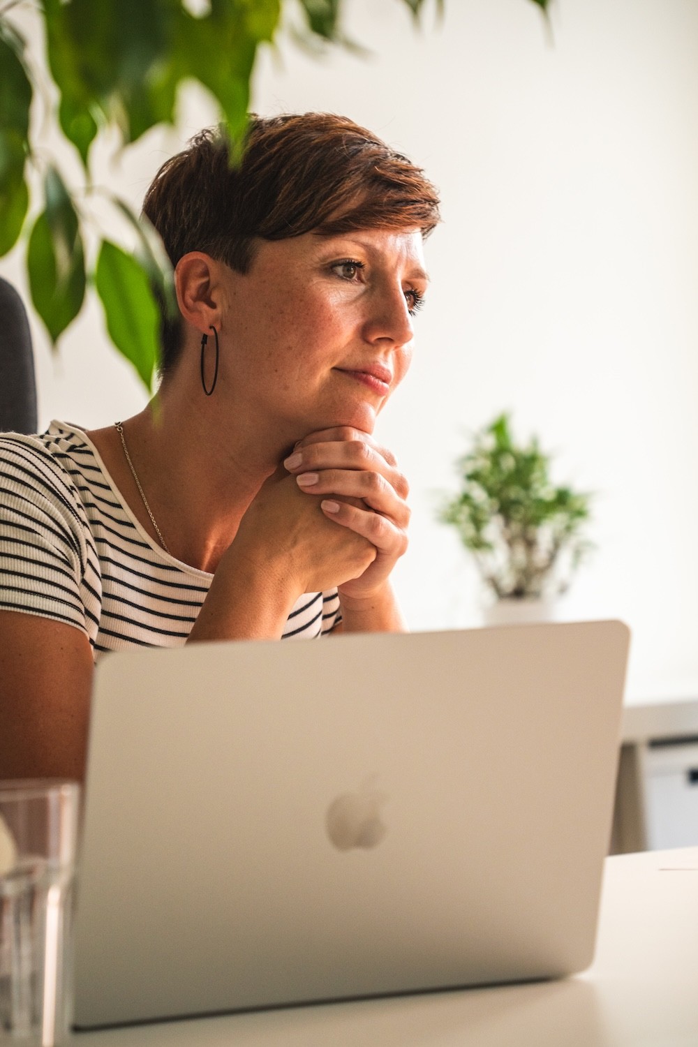 Person mit kurzem Haar und gestreiftem T-Shirt sitzt gespannt lächelnd vor einem Laptop mit Apfel-Logo in einem hellen Raum mit Pflanzen im Hintergrund