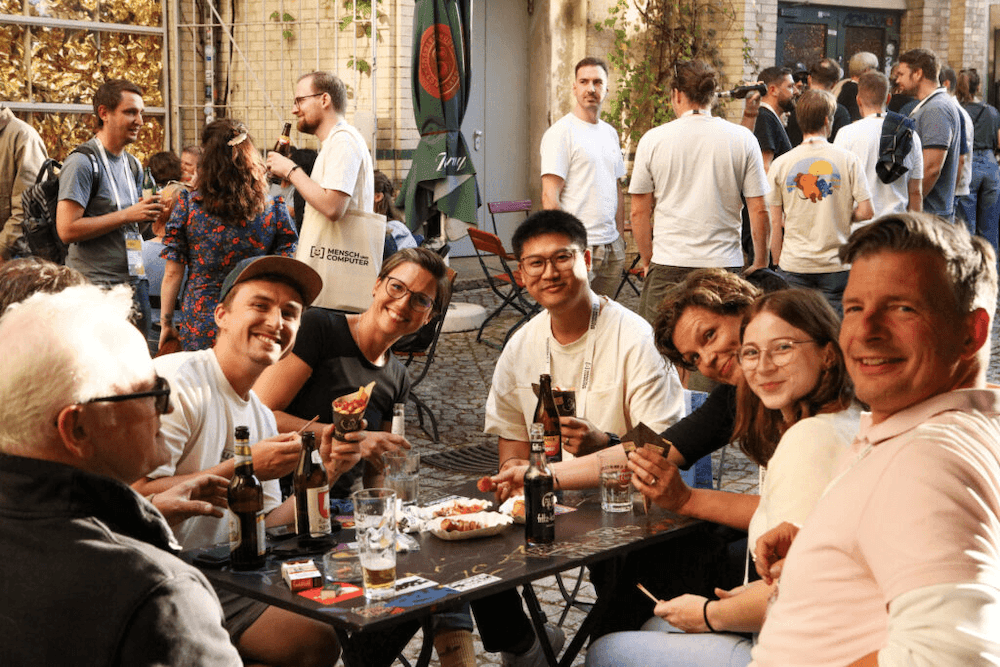 Gruppe von Menschen sitzt an einem Tisch im Freien und hält Getränke in der Hand, im Hintergrund weitere Personen in lockerer Atmosphäre vor einem Gebäude
