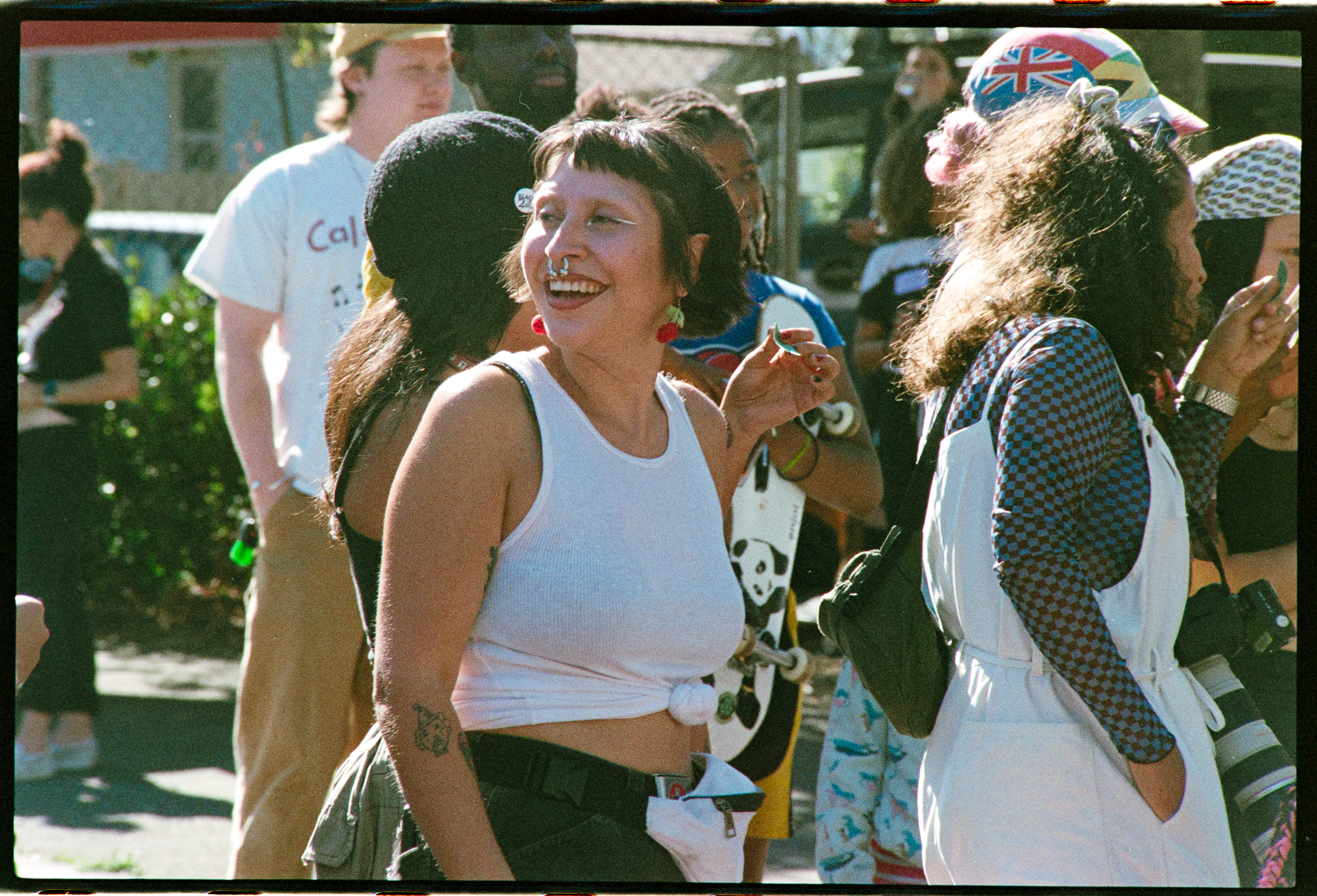 A group of people joyfully gather outdoors, featuring a woman in a white tank top smiling. The scene is lively, with colorful outfits and casual interactions.