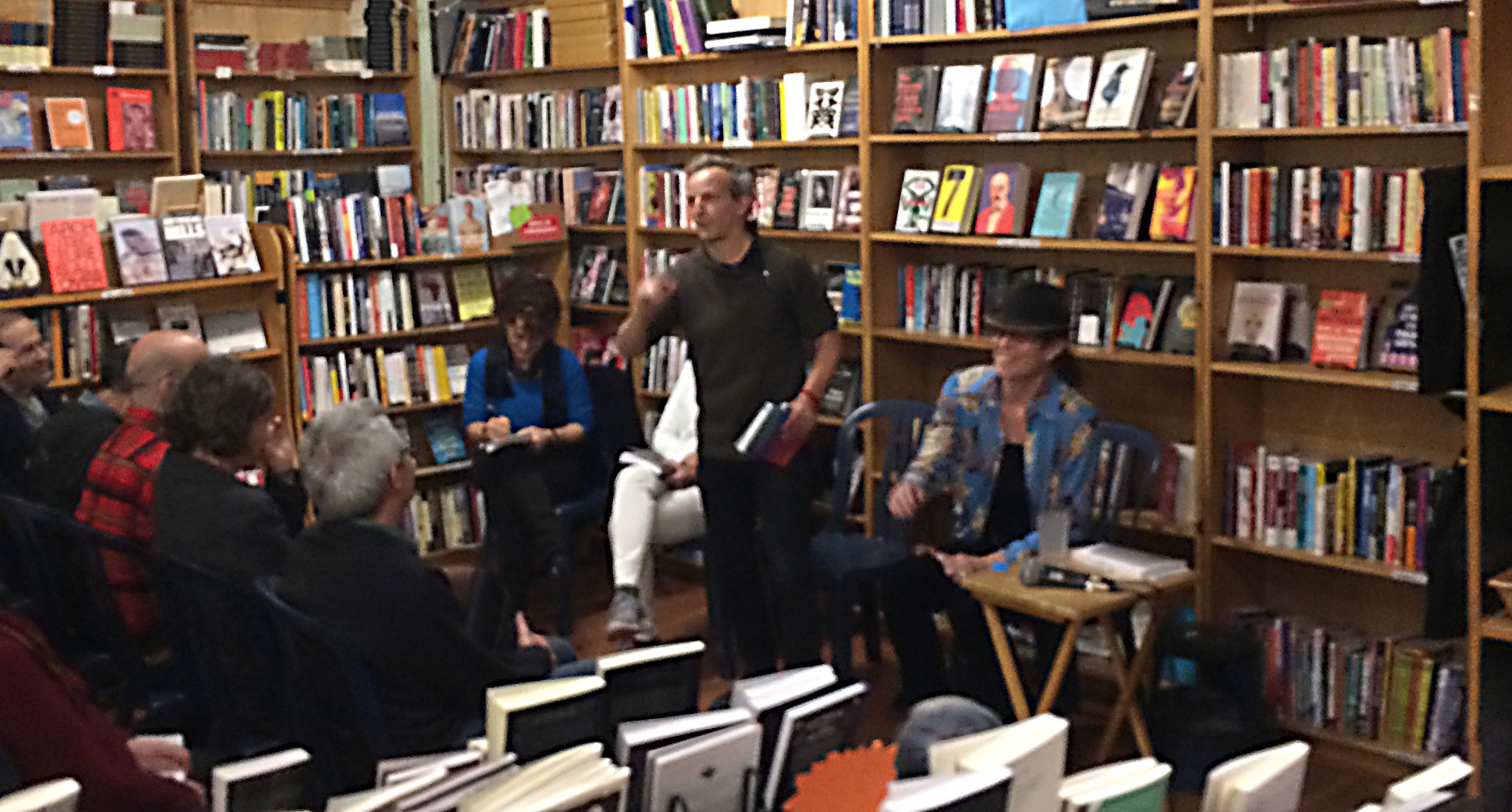 A photo of a group of people listening to a lecture while sitting in a library near some book shelfs.