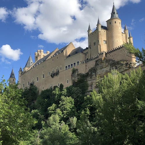 A large medieval castle with multiple turrets perched on a hilltop, surrounded by lush greenery and under a cloudy blue sky.