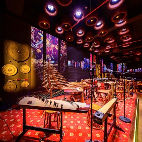 Sebuah bilik dipenuhi dengan alat muzik tradisional, seni dinding yang berwarna-warni, dan permaidani berwarna merah bercorak di bawah lampu siling bulat yang terang.